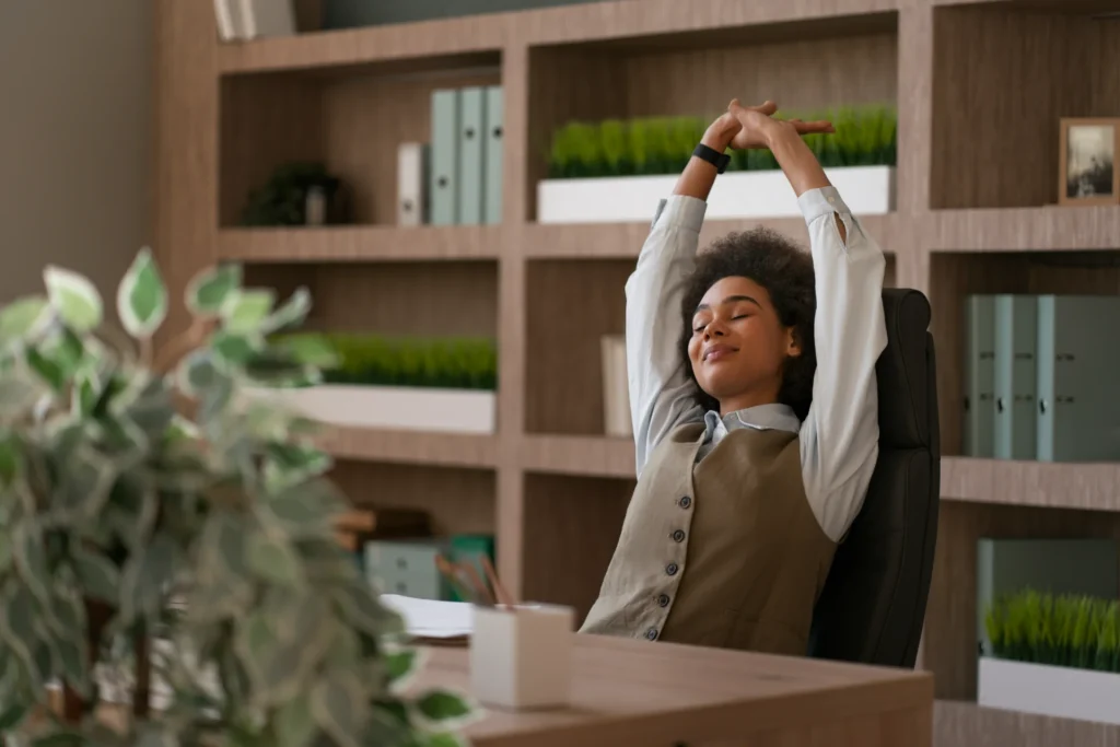Une femme aux cheveux bouclés s'étire les bras en souriant à son bureau avec une bibliothèque en arrière-plan