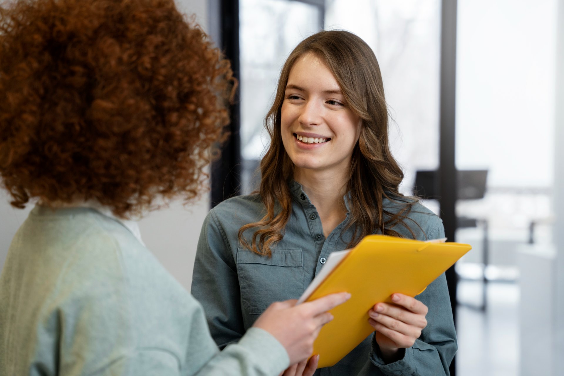 Deux collègues souriantes discutant dans un bureau, l'une tenant un dossier jaune lors d'un échange professionnel.